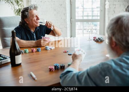 Jung im Herzen. Zwei glückliche reife Freunde spielen Karten und trinken Wein. Freuen Sie sich, aufgeregt. Kaukasische Männer spielen zu Hause. Aufrichtige Emotionen, Wohlbefinden, Gesichtsausdruck Konzept. Gutes Alter. Stockfoto