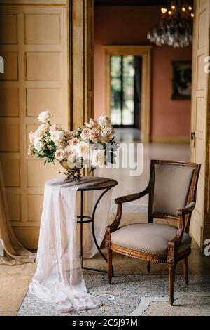 Bouquet von zarten cremefarbenen Rosen in Kupfervase auf dem Tisch im eleganten Raum mit Vintage-Sessel Stockfoto