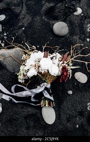 brautstrauß aus weißen Rosen, Lilie, Burgunder Nelken, protea, getrockneten Zweigen, Brunia und Leukadendron mit grauen und schwarzen Bändern auf dem schwarzen Sand in Stockfoto