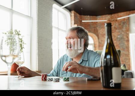 Aufregend. Glücklicher reifer Mann, der Karten spielt und Wein mit Freunden trinkt. Sieht begeistert aus, aufgeregt. Kaukasischer Mann spielt zu Hause. Aufrichtige Emotionen, Wohlbefinden, Gesichtsausdruck Konzept. Gutes Alter. Stockfoto