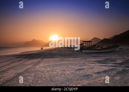 Sonnenuntergang mit blauem und orangefarbenem Himmel im Oman an der Küste von Dhofar und Salalah. Küstengebiet am Abend mit vielen Fischerbooten und Bergketten in Stockfoto