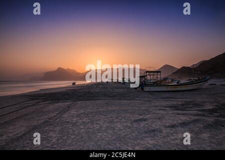 Sonnenuntergang mit blauem und orangefarbenem Himmel in Oman am Strand von Salalah Mughsail. Küstengebiet am Abend mit vielen Fischerbooten und Bergketten in der BA Stockfoto