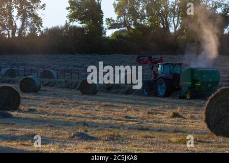 Hay Harvest Juni 2020 UK roter Traktor, Sonnenuntergang Ballenpressen das Heu. Große runde Ballen, für Pferdfutter Stockfoto