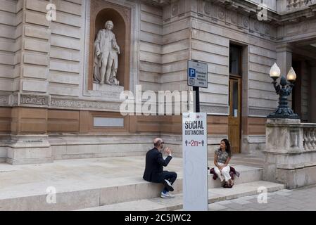Mit weiteren 89 britischen Opfer in den letzten 24 Stunden, die die Gesamtzahl der Opfer während der Coronavirus-Pandemie auf 43,995 bringen, werden Pubs, Restaurants, Friseure und einige Kunstgalerien wie hier, in der Royal Academy, wieder am Samstag, 4. Juli öffnen. Zwei Londoner sitzen und unterhalten sich nach den Regeln der Regierung, die auf vielen sozialen Fernanzeigen in der Hauptstadt am 2. Juli 2020 in London, England, gezeigt werden. Stockfoto