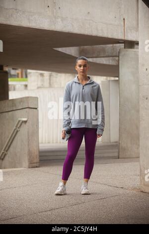 Eine Frau, die in violetten Leggings, grauem Sweatshirt und mit einer Getränkeflasche steht Stockfoto