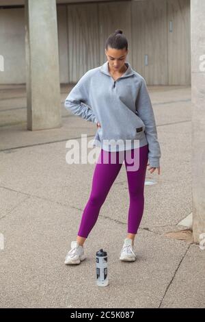 Eine Frau, die in violetten Leggings, grauem Sweatshirt und einer Trinkflasche auf dem Boden steht. Stockfoto