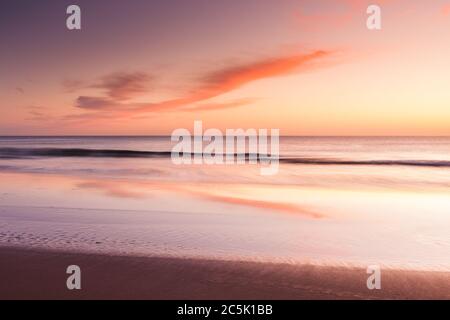 Eine ruhige, warme und farbenfrohe Aussicht über den Strand und die Bucht, die zum Horizont blickt und rollende Wellen auf den Strand waschen. Stockfoto