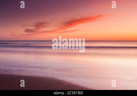 Eine ruhige, warme und farbenfrohe Aussicht über den Strand und die Bucht, die zum Horizont blickt und rollende Wellen auf den Strand waschen. Stockfoto