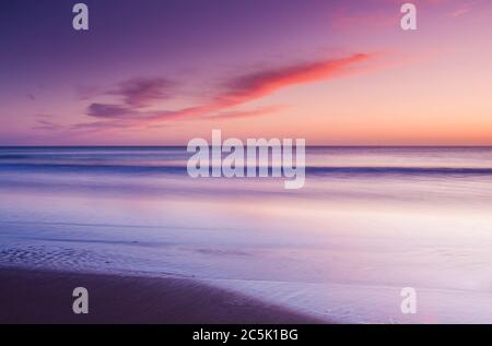 Eine ruhige, warme und farbenfrohe Aussicht über den Strand und die Bucht, die zum Horizont blickt und rollende Wellen auf den Strand waschen. Stockfoto