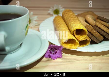 Waffelbrötchen und Kekse auf einem weißen Teller und eine Tasse Tee. Waffeln auf der Seite. Komposition auf einem hölzernen Hintergrund. Sichtbare Waffelstruktur Stockfoto