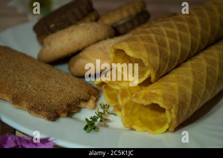 Waffelbrötchen und Kekse auf einem weißen Teller und eine Tasse Tee. Waffeln auf der Seite. Komposition auf einem hölzernen Hintergrund. Sichtbare Waffelstruktur Stockfoto