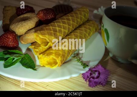 Waffelbrötchen und Kekse auf einem weißen Teller und eine Tasse Tee. Waffeln auf der Seite. Komposition auf einem hölzernen Hintergrund. Sichtbare Waffelstruktur Stockfoto