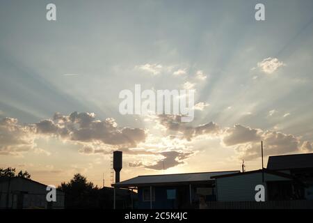 Sonnige Strahlen am Himmel. Zeit bei Sonnenuntergang oder Sonnenaufgang. Sonnenlicht bricht durch die Wolken am Himmel über den dunklen Silhouetten der Gebäude darunter Stockfoto