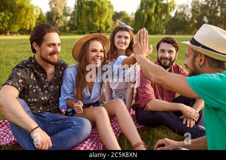 Gruppe von Freunden auf Gras sitzen, reden und High Fiving in der Landschaft. Fröhliche Menschen genießen Zeit zusammen im Freien im Park Stockfoto