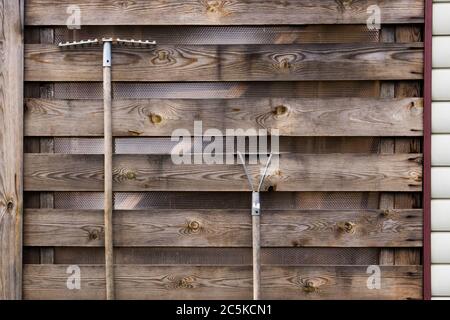 Rechen und Häcksler hängen an einer Holzwand von alten strukturierten Brettern. Stockfoto