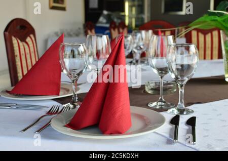 Restauranttisch gelegt und dekoriert, leere Gläser, Teller, Gabeln und Messer, rote Servietten und weiße Tischtücher Stockfoto