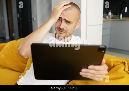Junge aufgeregt Kerl ist auf seinem Laptop surfen, zu Hause auf einem gemütlichen Sofa zu Hause sitzen. Finanzbörsen online. Schlechte Nachrichten Stockfoto