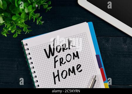Draufsicht auf offenes Notizbuch mit DER AUFSCHRIFT ARBEIT VON ZU HAUSE geschrieben. Grüne Blume, Tablett, Stift und Notizbuch auf Holzgrund. Stockfoto