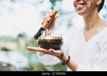 Junge lächelnde Frau spielt auf Messing tibetischen Klangschale draußen. Klangtherapie und Meditation Stockfoto