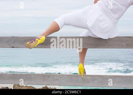 Attraktive Frau in weißen und gelben High Heel Schuhe Klettern auf hölzernen Handlauf, um Blick über den Ozean in verschwommenem Hintergrund und mit zu genießen Stockfoto