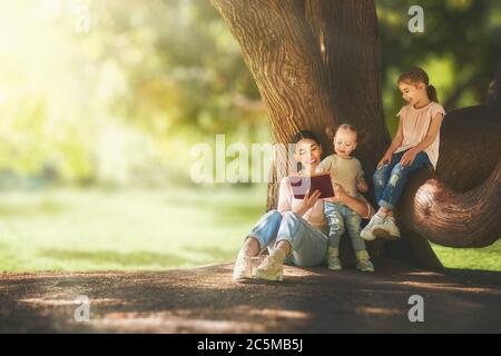 Mutter und Töchter sitzen unter dem Baum auf Sommerrasen. Glückliche Familie spielt draußen. Hübsche junge Mutter liest ihren Kindern im Park ein Buch Stockfoto