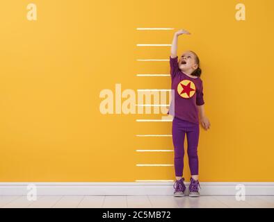 Kleines Kind spielt Superheld. Kid misst das Wachstum auf dem Hintergrund der hellen Farbe Wand. Mädchen Power Konzept. Gelb, rot und violett colo Stockfoto