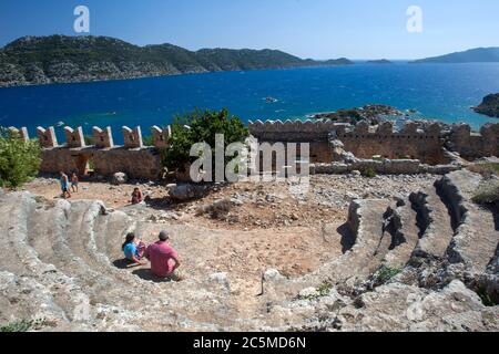 Touristen sitzen auf den Ruinen des antiken Theaters in der Festung Kreuzritter in Simena, die über der mediterranen Stadt Kalekoy in der Türkei sitzt. Stockfoto