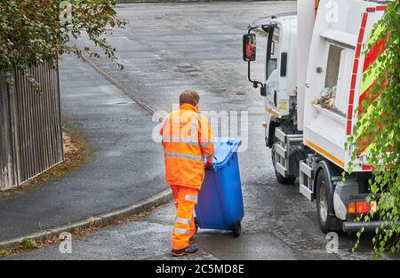 Ein Recycling-Sammler in orangefarbener Kleidung schiebt einen blauen Mülltonnen voller Haushaltsmüll in Richtung eines Müllsammelwagens. Stockfoto