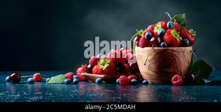 Beeren mit Blättern in Holzschale. Bunt gemischter Mix aus Erdbeeren, Blaubeeren, Himbeeren und Süßkirschen. Stockfoto