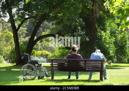 Ein älteres Paar mit Rollstuhl sitzt auf einer Bank im Park in Melbourne, Australien Stockfoto