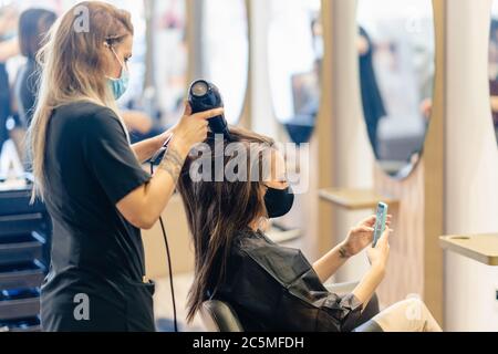 Friseur Trocknen der Haare ihrer Kunden mit einem Haartrockner tragen Schutzmasken in einem Beauty-Center. Stockfoto