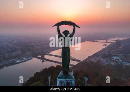 Luftaufnahme zur Freiheitsstatue mit Freiheitsbrücke und Donau im Hintergrund vom Gellert-Hügel bei Sonnenaufgang im Nebel in Budapest, Ungarn Stockfoto