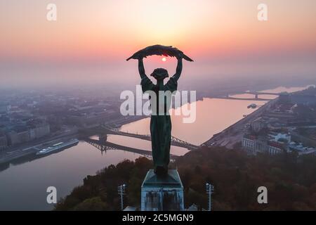 Luftaufnahme zur Freiheitsstatue mit Freiheitsbrücke und Donau im Hintergrund vom Gellert-Hügel bei Sonnenaufgang im Nebel in Budapest, Ungarn Stockfoto