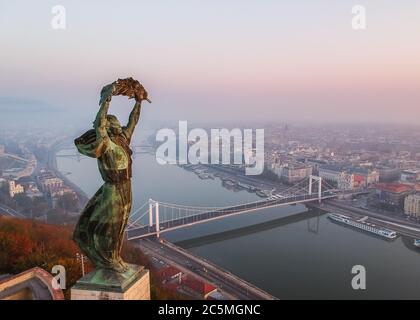 Luftaufnahme zur Freiheitsstatue mit Elisabeth Brücke und Donau vom Gellert Berg bei Sonnenaufgang im Nebel in Budapest, Ungarn. Stockfoto