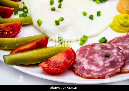 Leckeres Frühstück mit Eiern, Salami, Gurken, Kirschtomaten, Saucen und grünen Zwiebeln auf einem weißen Teller. Gesundes und leckeres Frühstück Stockfoto