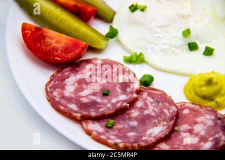 Leckeres Frühstück mit Eiern, Salami, Gurken, Kirschtomaten, Saucen und grünen Zwiebeln auf einem weißen Teller. Gesundes und leckeres Frühstück Stockfoto