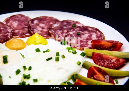 Leckeres Frühstück mit Eiern, Salami, Gurken, Kirschtomaten, Saucen und grünen Zwiebeln auf einem weißen Teller. Gesundes und leckeres Frühstück Stockfoto