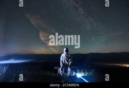 Beleuchteter Astronaut in weißem Raumanzug und Helm, der nachts auf der Bank auf dem Hügel sitzt. Bergrücken und Himmel voller Sterne auf dem Hintergrund. Konzept der Raumfahrt, Galaxie und Kosmonauten. Stockfoto