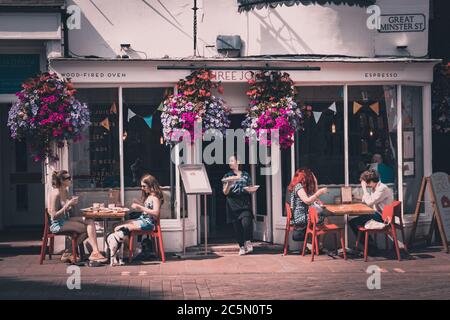 Die Vorderseite eines Cafés mit Leuten, die an Tischen sitzen In der Sonne genießen Essen und einen Kaffee mit einem Kellnerin trägt Teller mit Essen aus der Tür Stockfoto