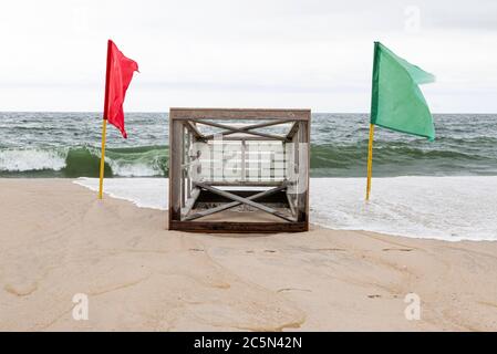 Ein Rettungsschwimmer steht auf dem Sand am Strand mit den Wellen im Hintergrund und roten und grünen Fahnen auf beiden Seiten, während das Wasser hinüberrollt. Stockfoto