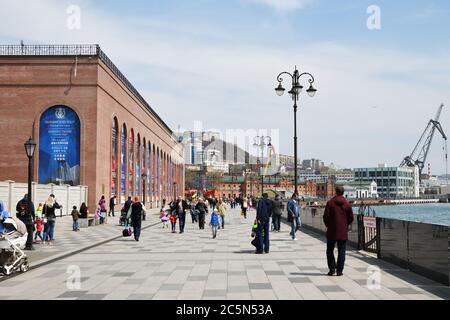 Wladiwostok, Russland - 28. April 2019: Die Menschen gehen entlang der Promenade Korabelnaya Naberezhnaya, Frachthafen im Hintergrund Stockfoto