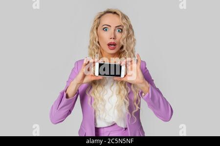 Porträt der erstaunt blonde Frau eindrucksvoll zeigt Smartphone-Bildschirm, halten Handy horizontal, amüsant offenen Mund Stockfoto