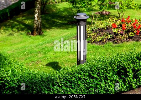 In den Büschen zwischen der Landschaft ist eine eiserne Gartenlaterne mit Blumenbeeten und grünem Gras an einem sonnigen Sommertag in einem gepflegten Garten installiert. Stockfoto