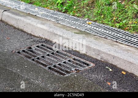 Kanalschachtgitter auf einer nassen Asphaltstraße nach Regen in der Nähe der Bordsteinkante im Hintergrund hinter der Bordsteinkante ein Abflusskanal mit einem Gitter bedeckt. Stockfoto