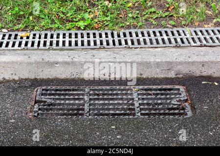 Kanalschachtgitter auf einer Asphaltstraße in der Nähe der Bordsteinkante im Hintergrund hinter der Bordsteinkante ein Abflusskanal mit einem Grill bedeckt. Stockfoto