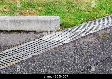 Eisen-Gitter-Entwässerungssystem auf einer asphaltierten Straße mit einem Bordstein auf der Seite der Straße grünes Gras., Nahaufnahme. Stockfoto