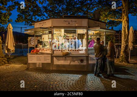 Der Imbiss Riviera Lebensmittelladen am Ufer des Zürichsees serviert abends unter anderem Cervelat, die Nationalwurst der Schweiz, Zürich, Schweiz. Kunden warten an der beleuchteten Theke, während Bäume den gepflasterten Platz säumen. Stockfoto