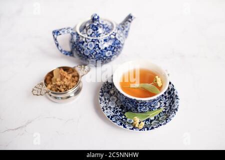 Traditionelle Teetasse mit Lindentee und Lindenblume auf einem weißen Marmortisch mit passender Teekanne im Hintergrund Stockfoto