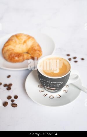 Kaffeetasse mit Löffel und Kaffeebohnen neben einem leckeren Croissant auf einem weißen Marmortisch Stockfoto