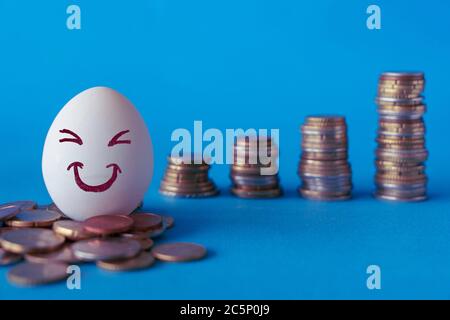 Gesichter auf den Eiern, Investitionskonzept. Glücklicher Investor lächelt vor seinem wachsenden Geld. Blauer Hintergrund mit Münzstapeln Stockfoto